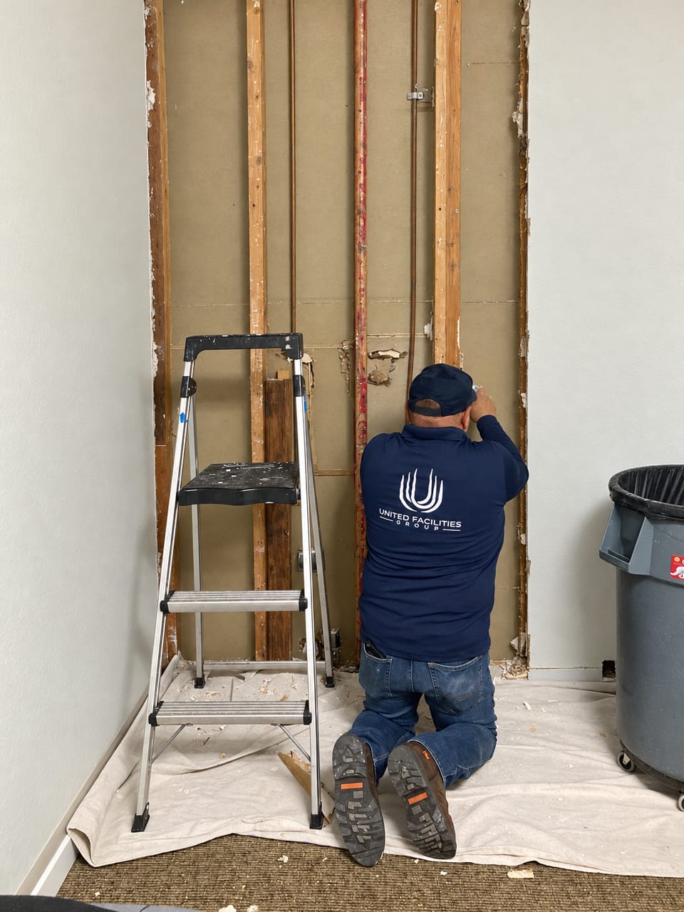 A UFG technician in a navy UFG shirt kneels at a wall opened to its studs, with a step ladder and trash can beside him