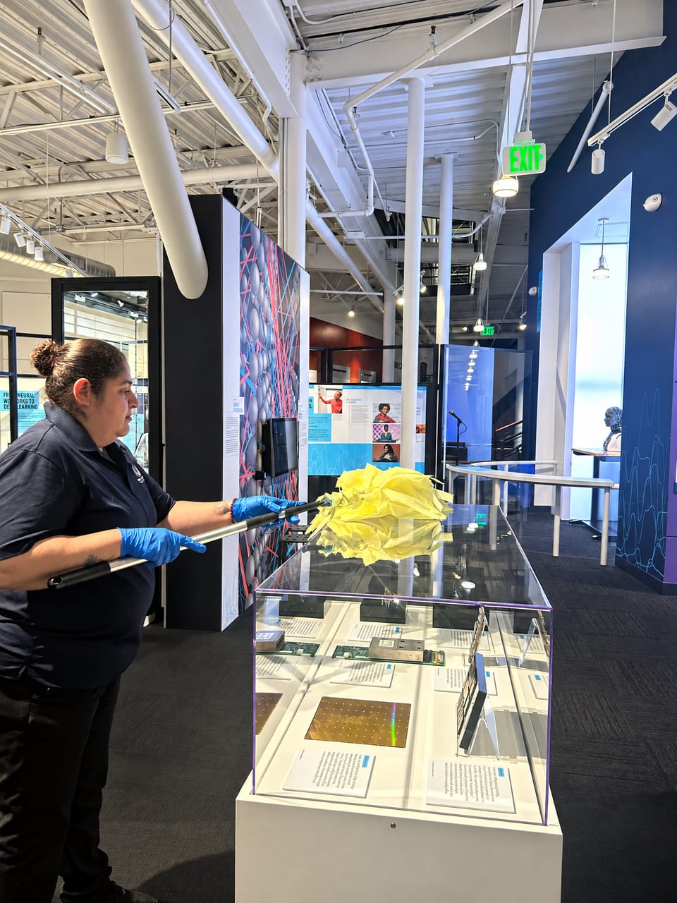 A UFG team member in blue gloves dusts a glass display case in an exhibit hall with a long yellow duster
