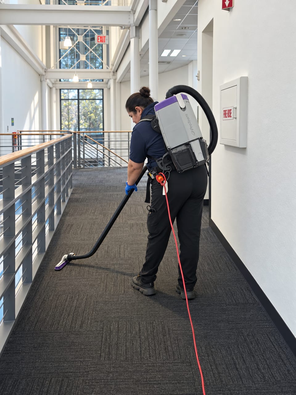 A UFG team member in a backpack vacuum, seen from behind, cleans dark carpet on a museum walkway, a red cord trailing