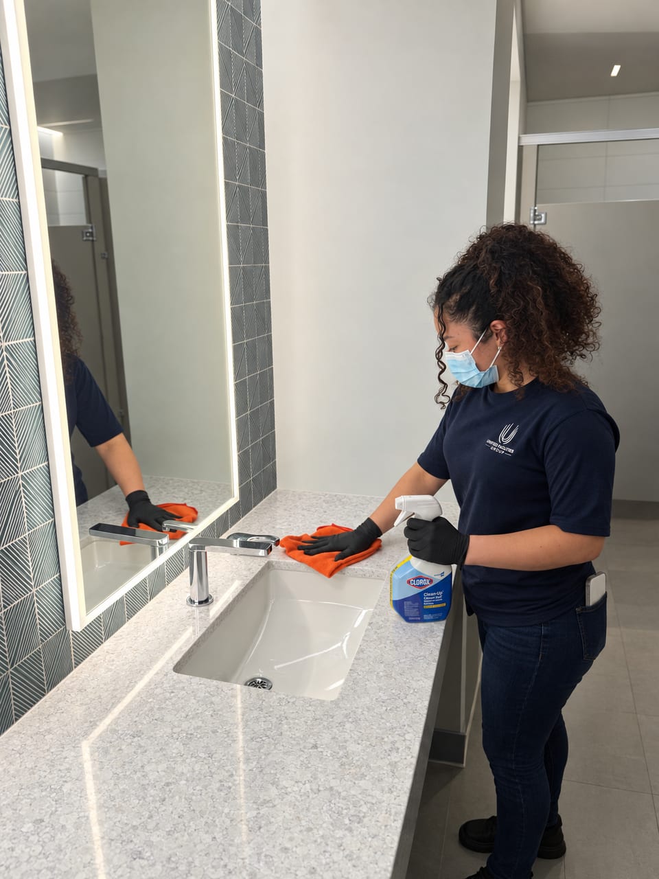 A UFG team member in a mask and black gloves wipes the counter beside a restroom sink with an orange cloth