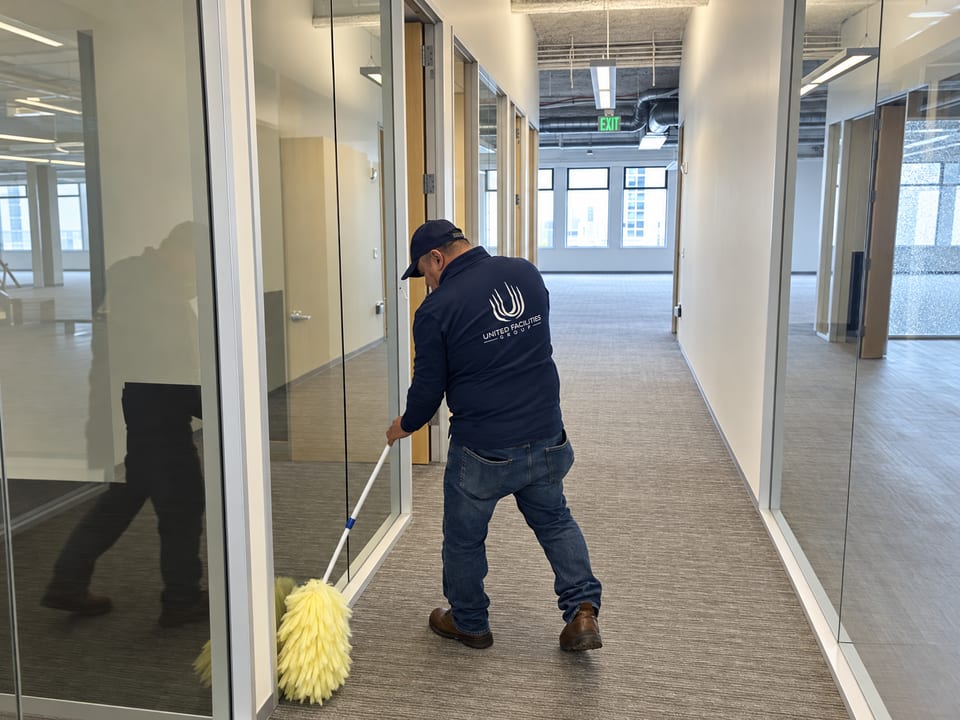 A UFG team member in a navy UFG jacket and cap dusts the base of glass partitions in a corridor with a long yellow duster