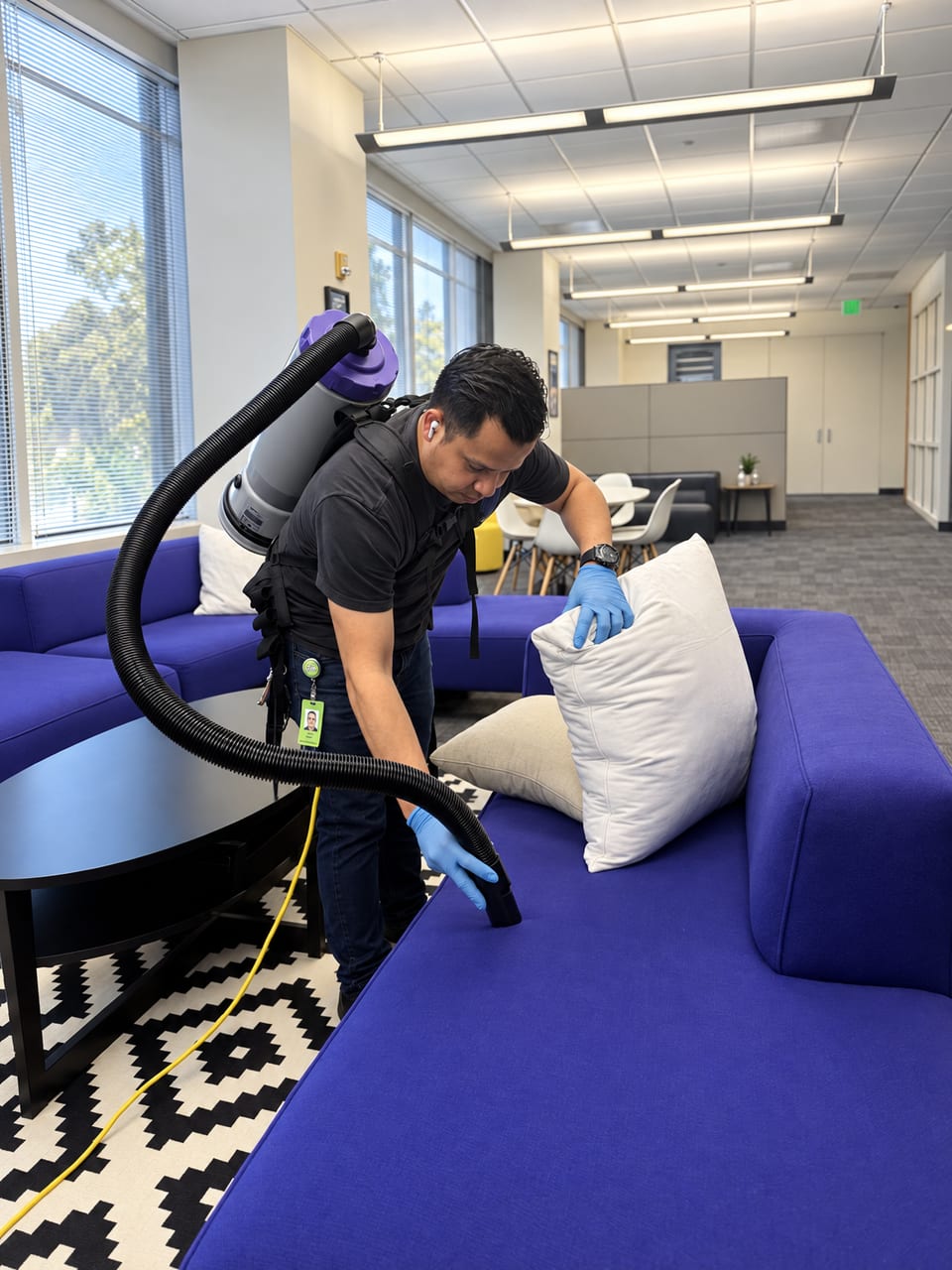 A UFG team member in blue gloves vacuums a purple office sofa with a backpack vacuum, holding a cushion aside