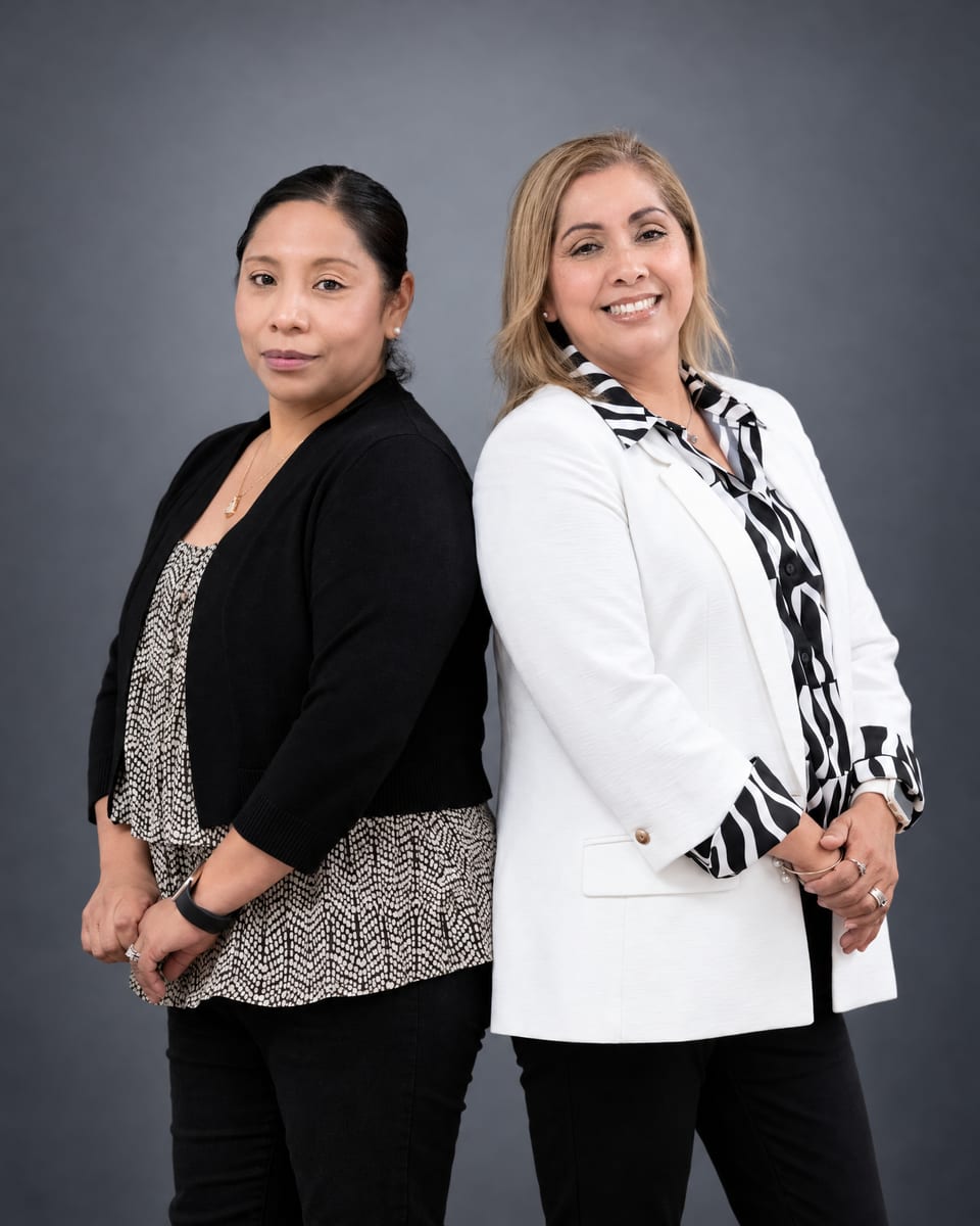 Two women stand back to back with hands clasped, one in a black cardigan over a print top, one in a white blazer and scarf