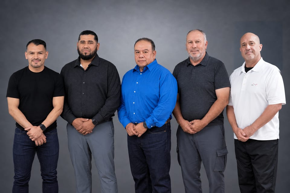 Five men standing in a row in black, grey, blue and white shirts against a grey studio backdrop.