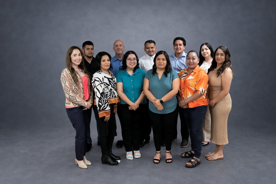 Eleven team members pose close together in teal, orange, leaf-print, light-blue and beige tops on a grey backdrop