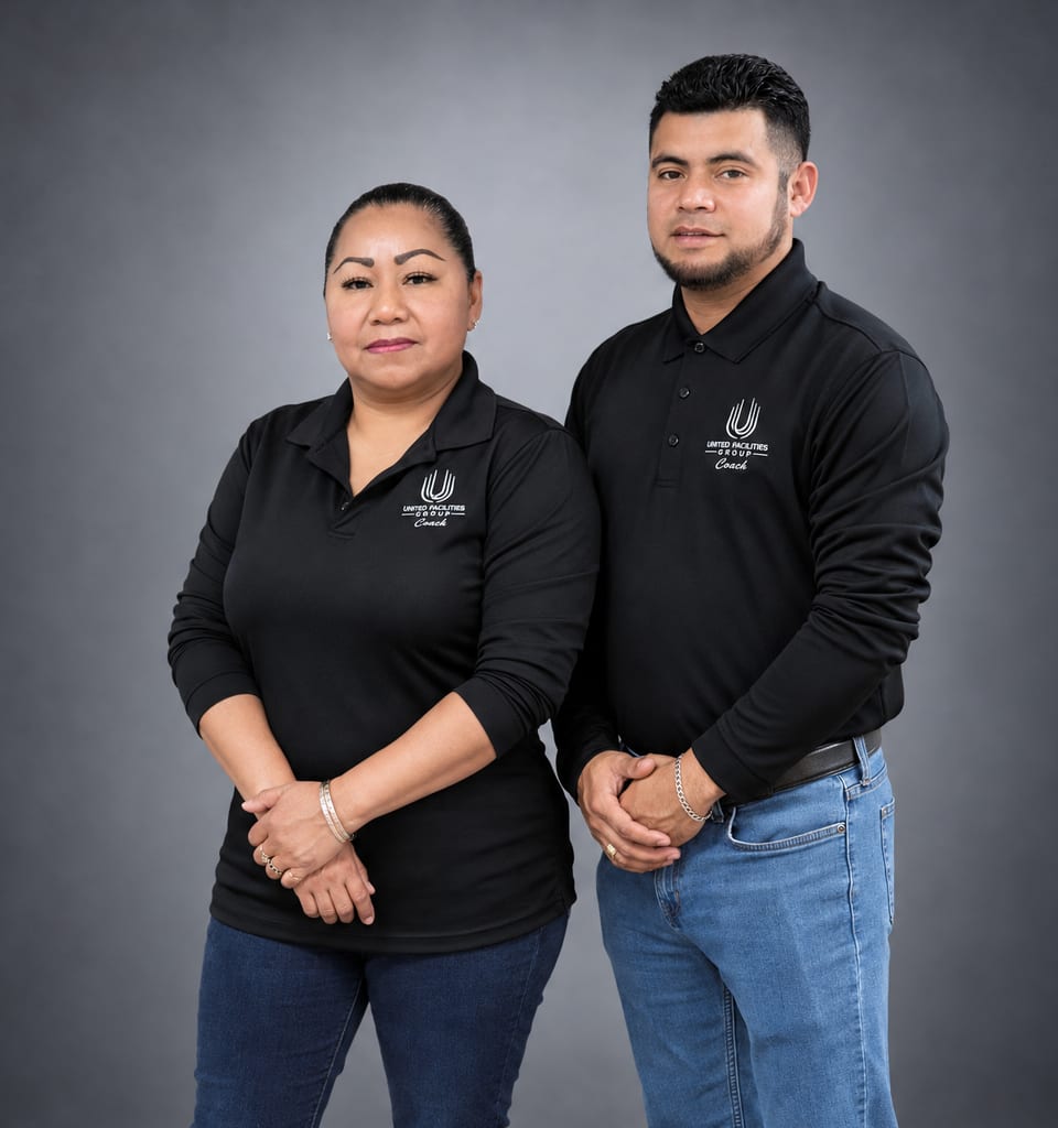 A woman and a man in black UFG Coach polos stand side by side, hands clasped, on a grey backdrop