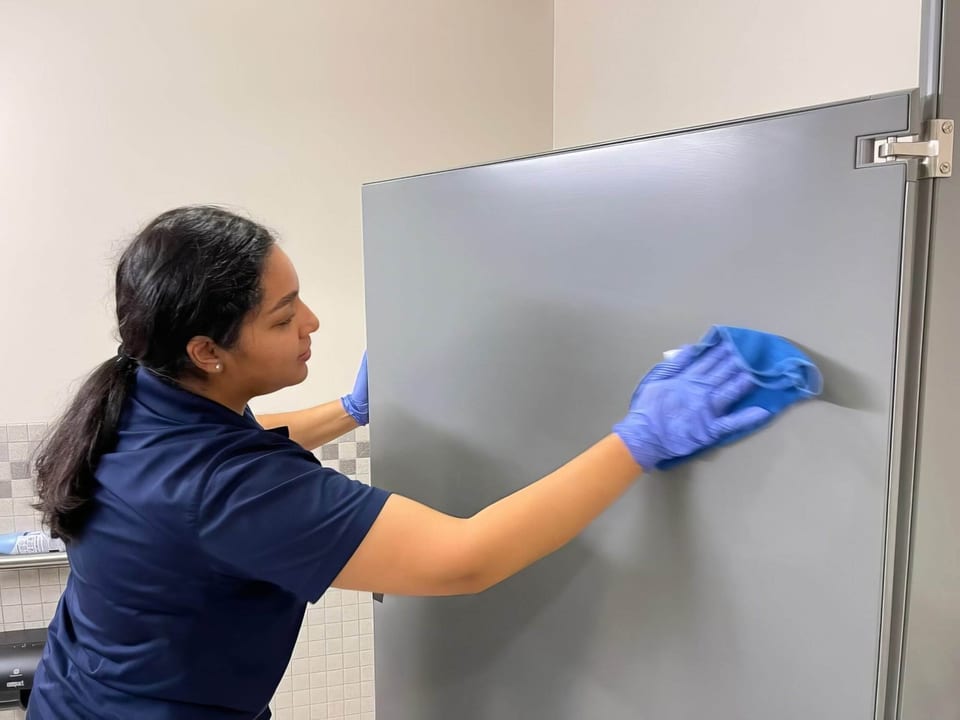 A UFG team member in a navy polo and purple gloves wipes a grey restroom stall door with a blue cloth