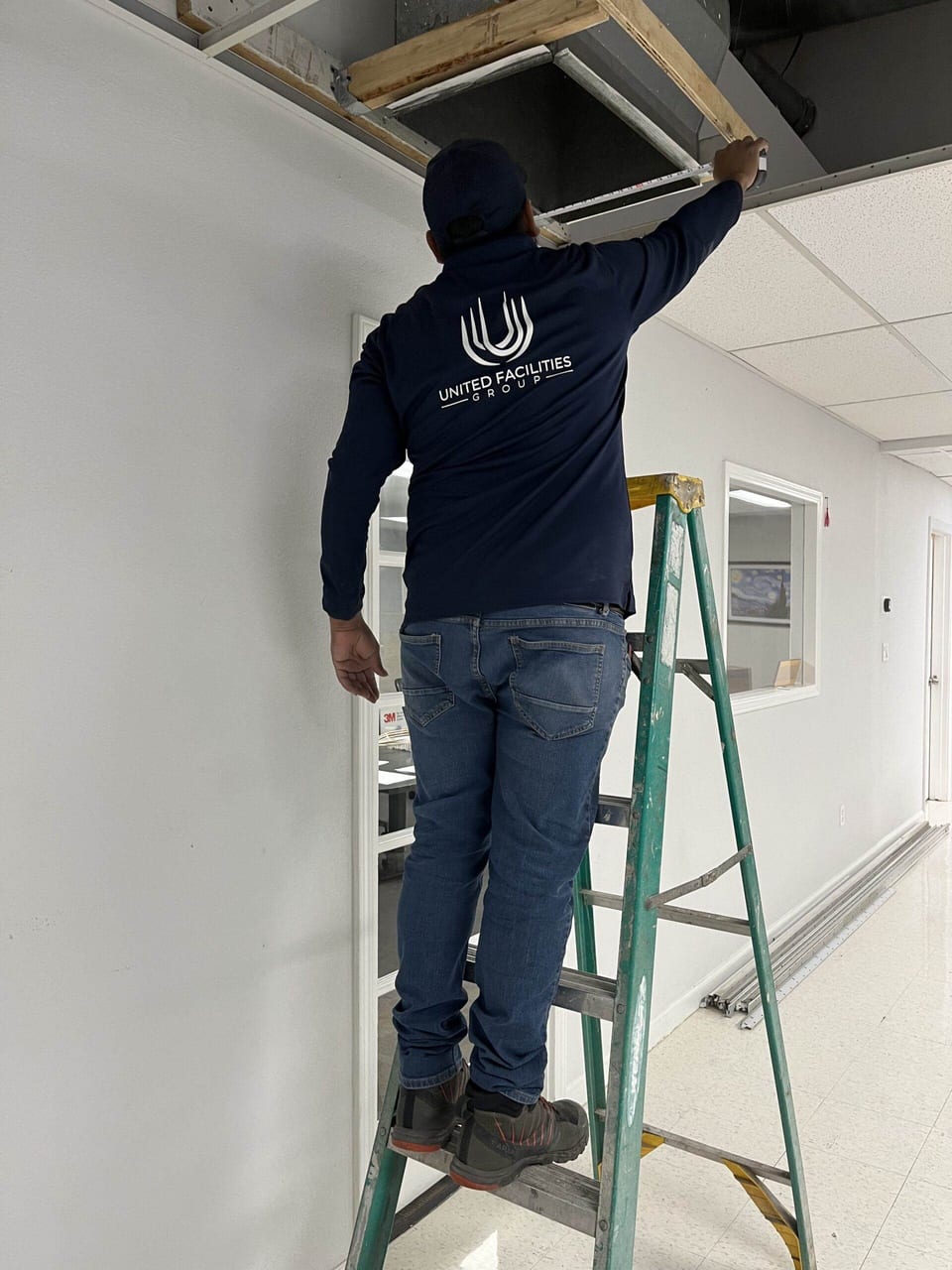 A UFG technician in a navy UFG shirt stands on a green ladder and reaches into an opened ceiling access panel