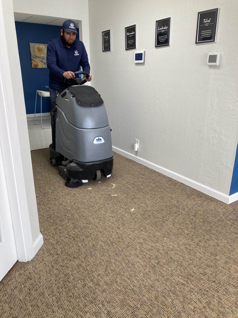 A UFG team member in a navy cap and UFG pullover rides a grey floor scrubber across office carpet beside framed plaques
