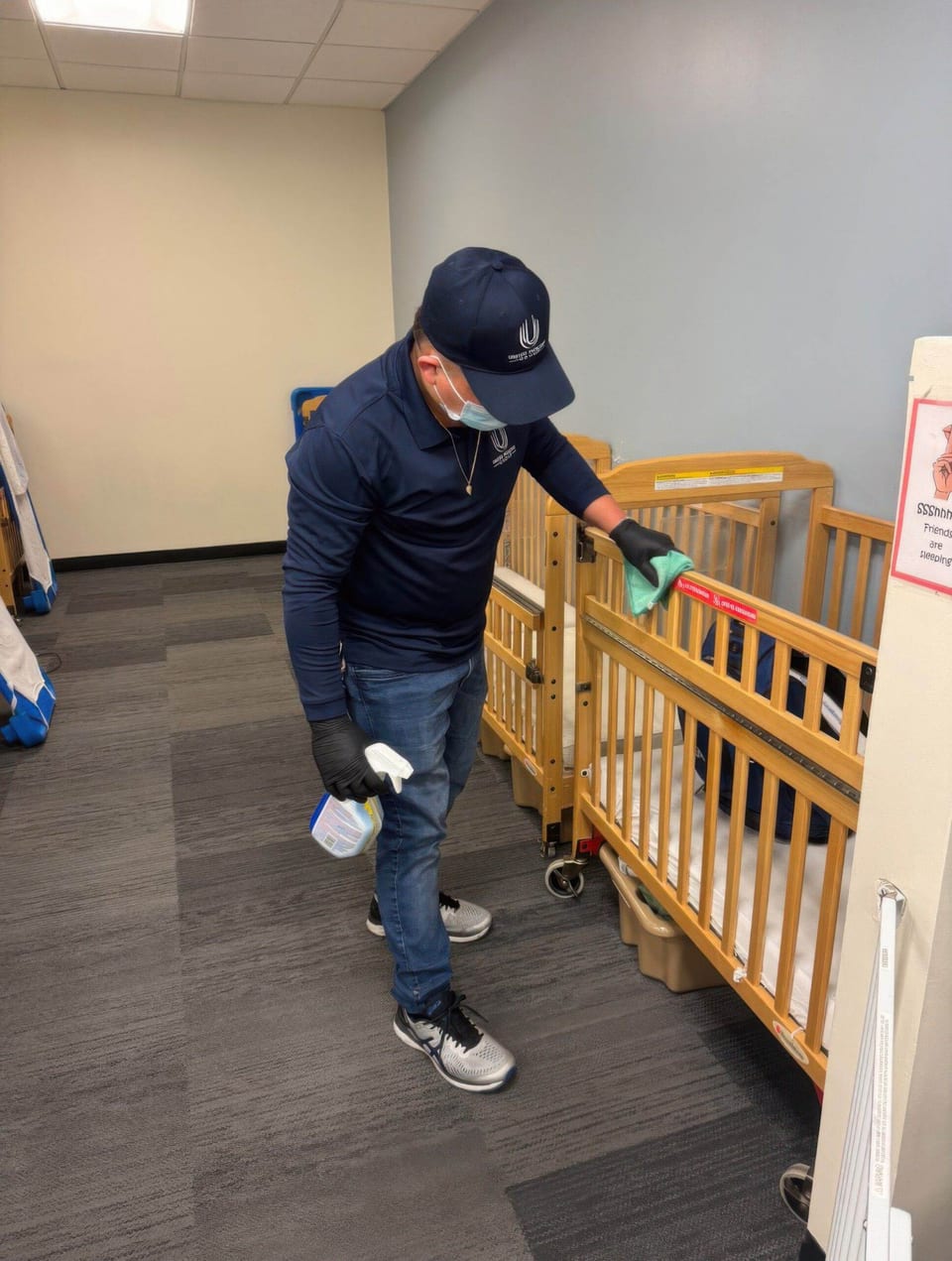 A UFG team member in a navy cap, mask and black gloves wipes a wooden crib rail in a nap room, spray bottle in hand