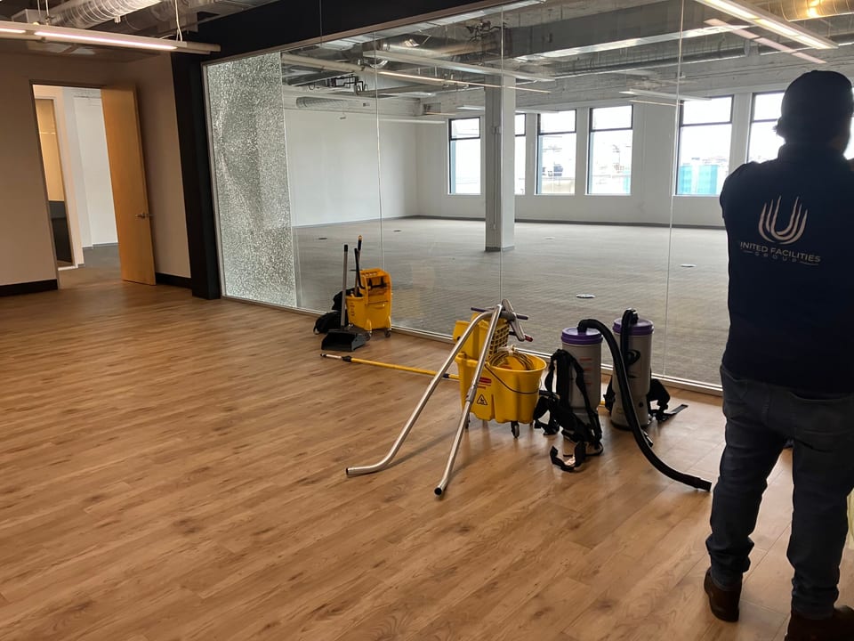 A UFG team member in a navy UFG shirt stands in a large empty glass-walled office beside mop buckets and backpack vacuums