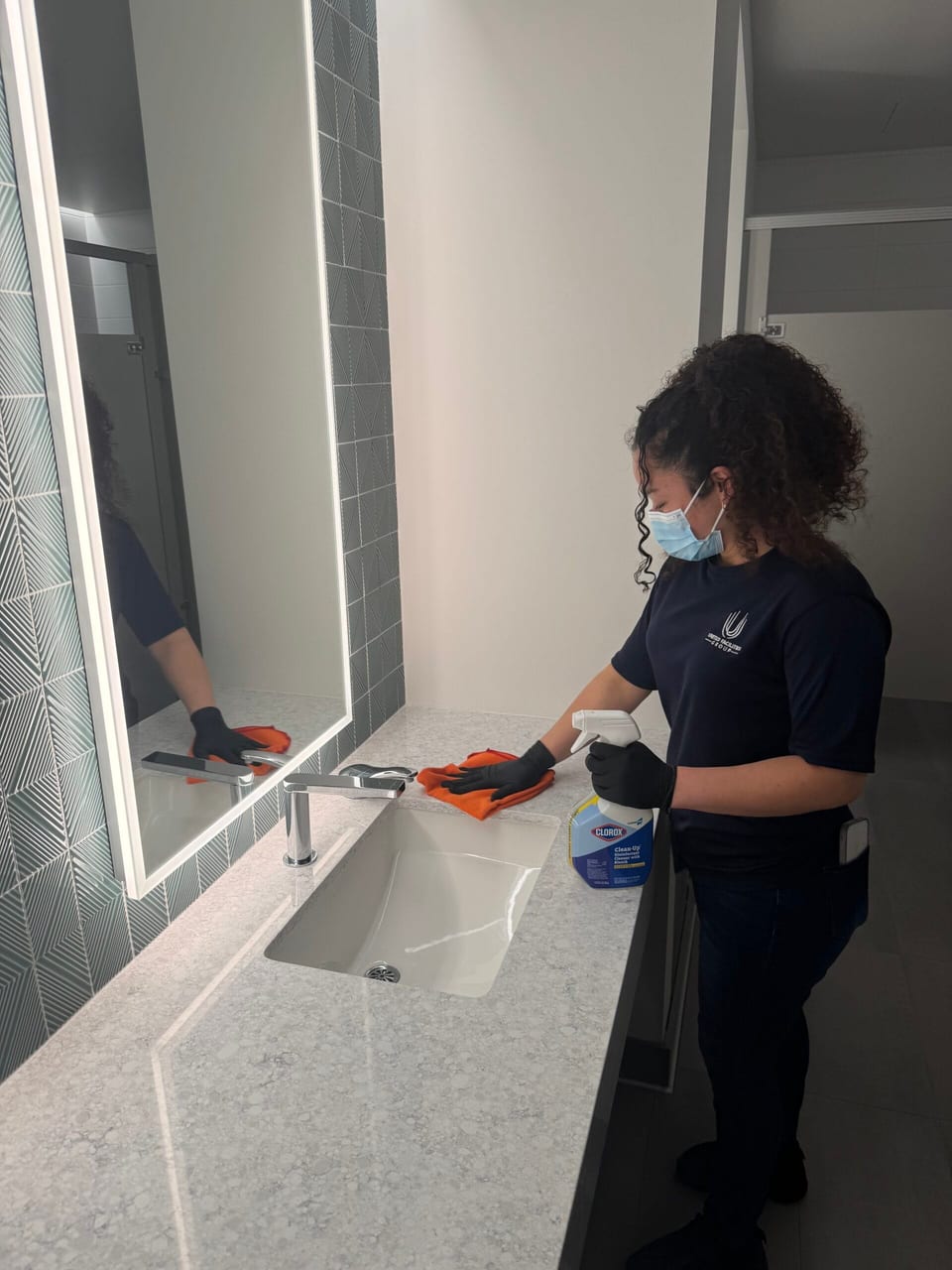 A UFG team member in a mask and black gloves wipes a restroom vanity with an orange cloth, holding a disinfectant spray