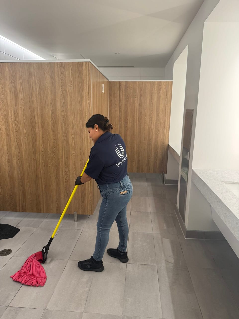A UFG team member in a navy UFG polo mops a restroom's tile floor with a red mop beside wood-panel stalls