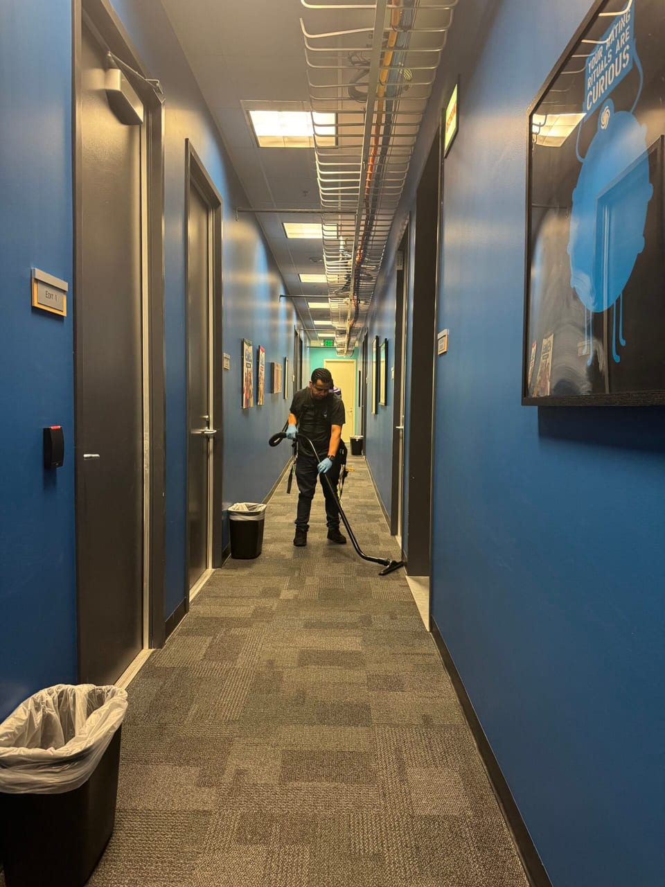 A UFG team member with a backpack vacuum cleans the carpet of a long blue-walled office corridor