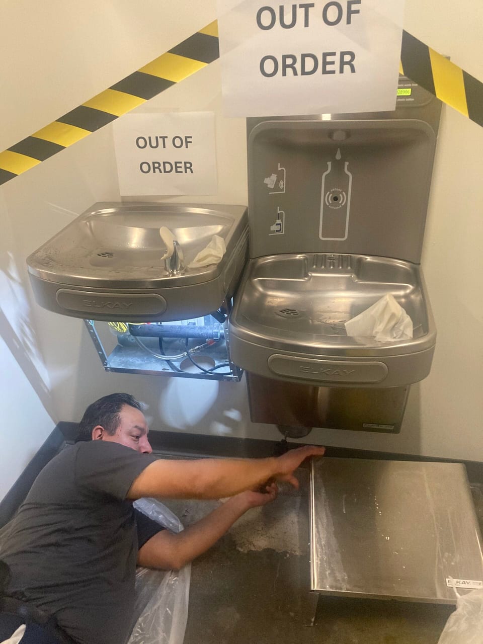 A UFG technician lies on the floor reaching under two wall-mounted drinking fountains to repair their plumbing