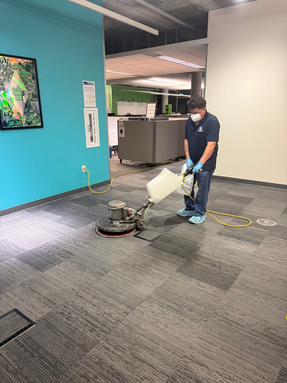 A UFG team member in a mask, gloves and shoe covers runs a rotary floor machine across office carpet beside a teal wall