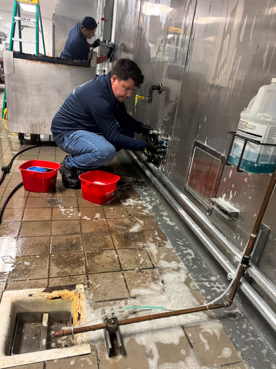 Two UFG team members in navy pullovers work in a commercial kitchen, one crouched scrubbing wall pipe valves with a sponge