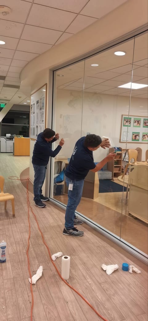 Two UFG team members in navy shirts wipe down a curved glass wall with paper towels in a childcare centre