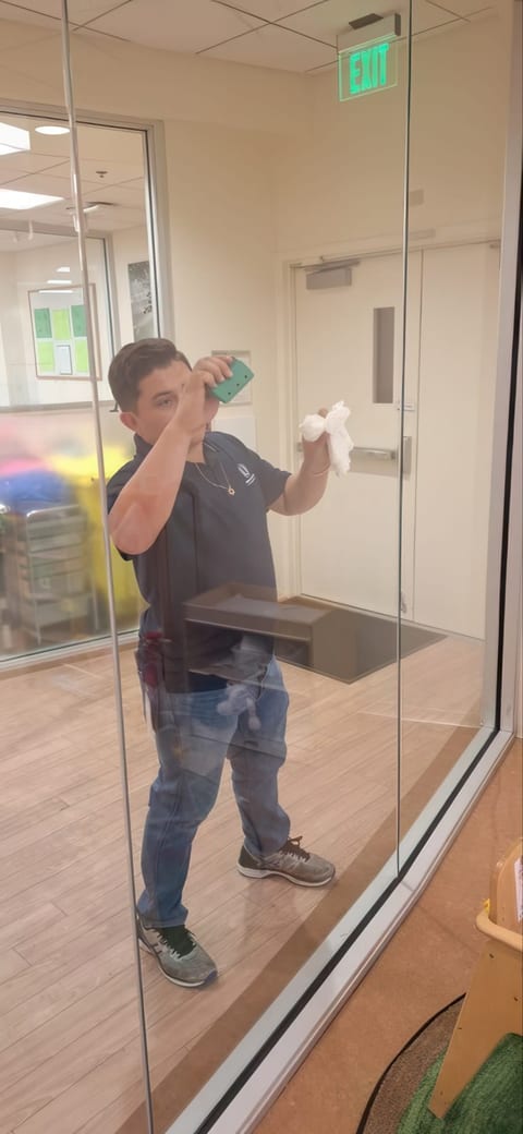 A UFG team member sprays and wipes a glass wall with a paper towel, seen through the glass beneath an exit sign