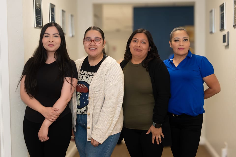Four women stand together in an office hallway in a black top, a cream cardigan, an olive top and a bright blue polo