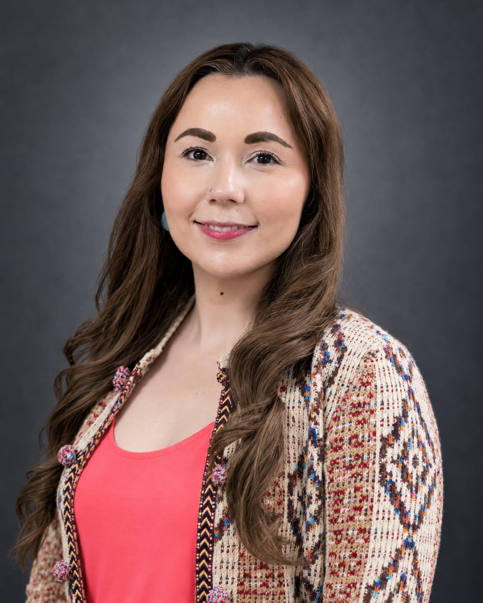Head-and-shoulders portrait of a woman with long wavy brown hair in a patterned cardigan over a coral top on a grey backdrop